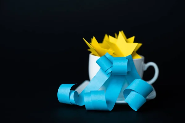 white cup with paper stars near blue ribbons isolated on black 