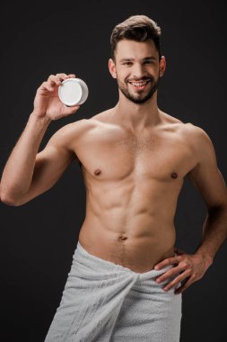 Havluyla gülümseyen yakışıklı adam elinde plastik kapla yüz kremi ile siyah üzerine izole edilmiş.
