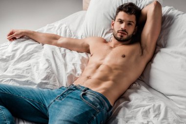 Kot pantolonlu, yakışıklı, seksi, üstsüz adam gri yatakta yatıyor.