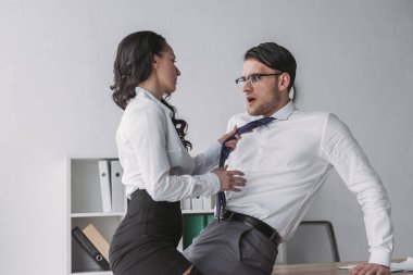 Seksi iş kadını, ofiste onu baştan çıkarırken şok olmuş iş adamının kravatına dokunuyor.