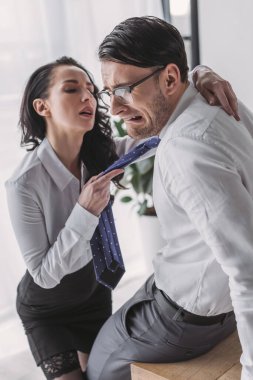 Seksi sekreter kravatını tutuyor ve ofiste onu baştan çıkarırken korkmuş erkek arkadaşına sarılıyor.