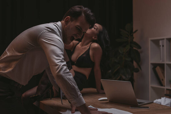 angry businessman screaming while standing ner papers while sexy secretary sitting on desk in lingerie