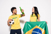 multicultural couple of happy football fans holding brazilian flag, ball and bottle of beer on grey