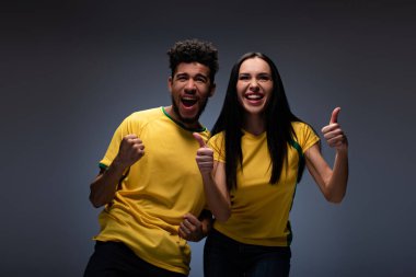 cheerful multiethnic couple of football fans in yellow t-shirts showing thumbs up on grey clipart