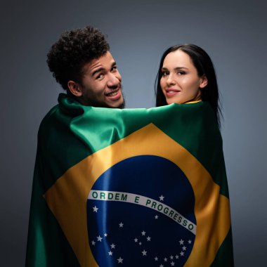 multicultural couple of positive football fans with brazil flag on grey clipart