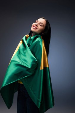 happy female football fan wrapped in brazilian flag on grey clipart