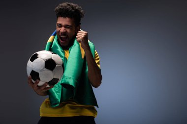 Brezilya bayraklı neşeli Afrikalı Amerikalı adam bağırıyor ve futbol topunu gri üzerinde tutuyor.