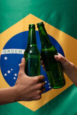 cropped view of couple of football fans clinking with bottles of beer with brazil flag behind clipart