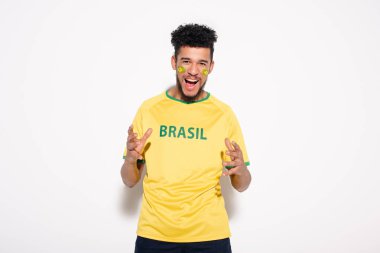 emotional african american football fan in yellow t-shirt with brazil sign gesturing and shouting on grey clipart