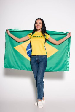 Çekici mutlu bayan futbol fanatiği gri üzerinde Brezilya bayrağı tutuyor