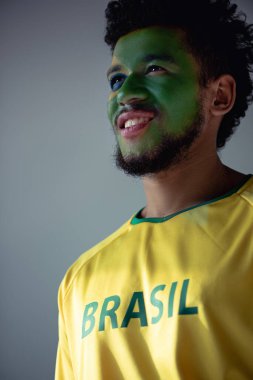 positive african american football fan with face painted as brazilian flag isolated on grey clipart
