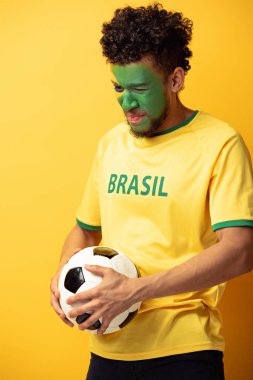 Vurgulu bir Afrikalı Amerikan futbol fanatiği. Yüzü Brezilya bayrağı gibi boyanmış. Sarı topun üzerinde.