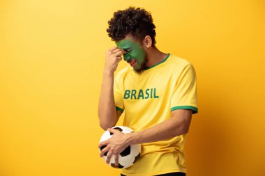 Kafası karışmış Afrikalı Amerikan futbol fanatiği. Yüzü Brezilya bayrağı şeklinde boyanmış. Sarı topun üzerinde.