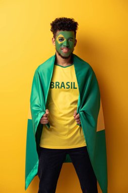 happy african american football fan with painted face wrapped in brazilian flag on yellow clipart