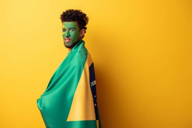 handsome african american football fan with painted face wrapped in brazilian flag on yellow clipart