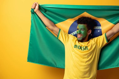 happy african american football fan with painted face holding brazilian flag on yellow clipart