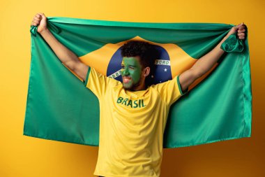 cheerful african american football fan with painted face holding brazilian flag on yellow clipart