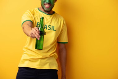Yüzü Brezilya bayraklarına boyanmış, sarı bira şişesi taşıyan Afro-Amerikan futbol taraftarı görüntüsü.