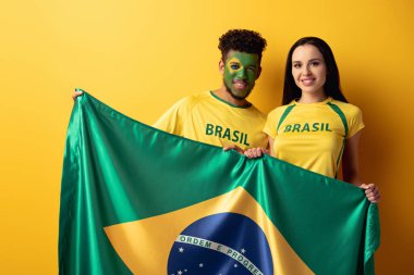 Erkek Afro-Amerikan futbol fanatiği, yüzü boyalı ve mutlu bir kız, sarı bayrakla Brezilya bayrağı taşıyor. 