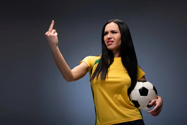 beautiful aggressive football fan holding ball while showing middle finger on grey