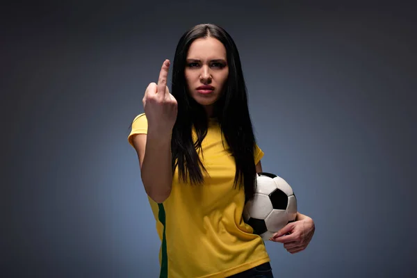 angry female football fan holding ball while showing middle finger on grey