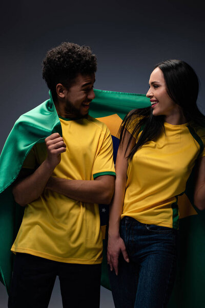 пара мультиэтнических улыбающихся футбольных болельщиков с бразильским флагом на сером
