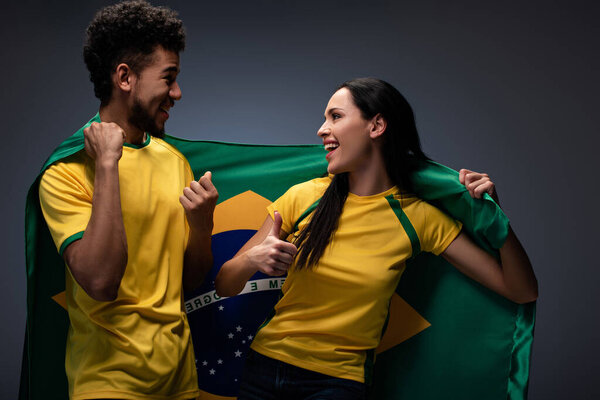 Мультикультурная пара восторженных футбольных болельщиков с бразильским флагом на сером
