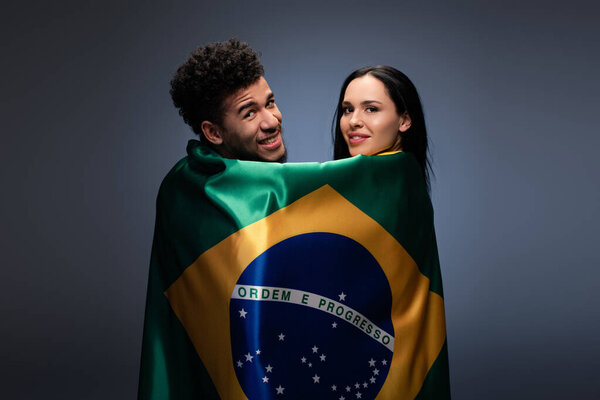 мультикультурная пара улыбающихся футбольных болельщиков с бразильским флагом на сером
