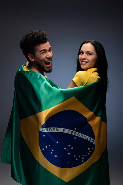 мультикультурная пара улыбающихся футбольных болельщиков с бразильским флагом на сером
