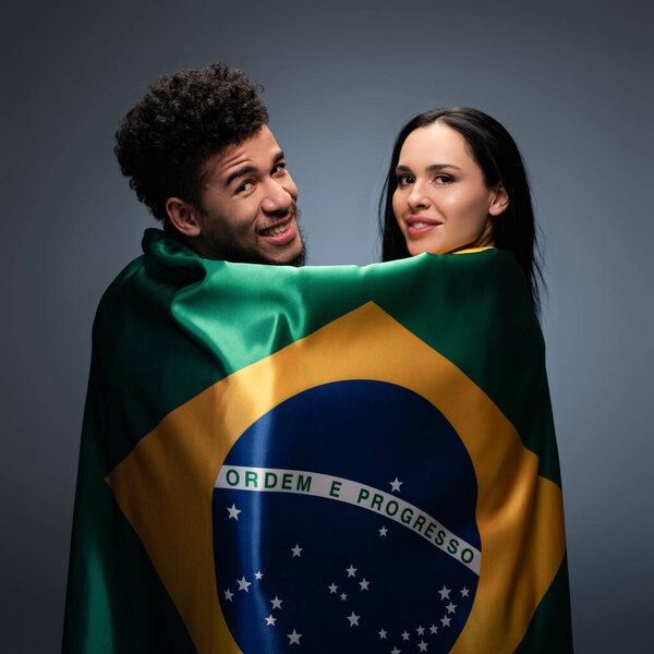 Мультикультурная пара позитивных футбольных болельщиков с бразильским флагом на сером
