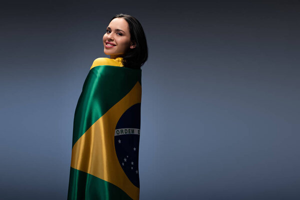 красивая веселая футбольная фанатка, завернутая в бразильский флаг на сером
