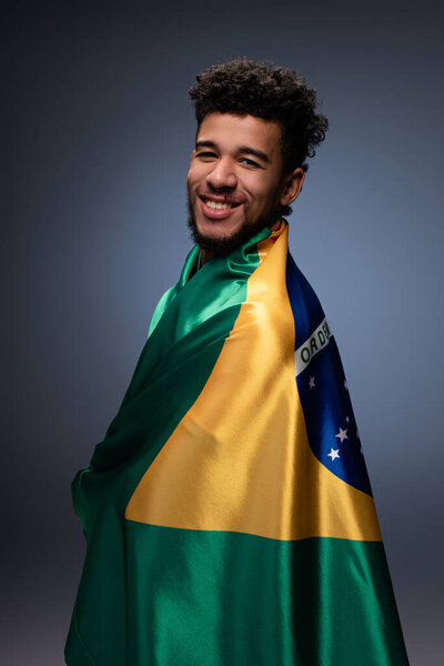улыбающийся африканский футбольный болельщик, завернутый в бразильский флаг на сером
