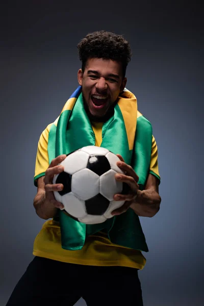 emotional african american man with brazilian flag screaming and holding football ball on grey