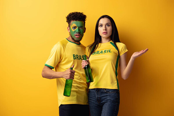 Африканский футбольный фанат мужского пола с раскрашенным лицом и смущенной девушкой, держащей бутылки пива на желтом
 
