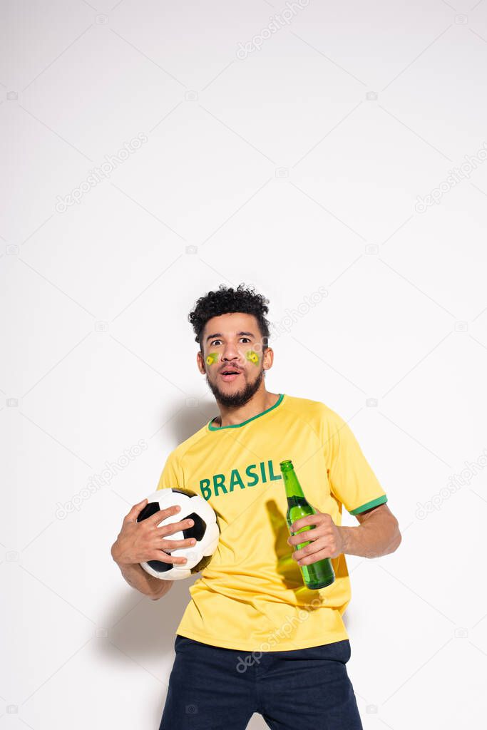 shocked african american man holding football ball and bottle of beer on grey