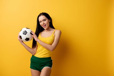 Güzel, neşeli bayan futbol fanatiği göz kırpıyor ve sarıda topu tutuyor.