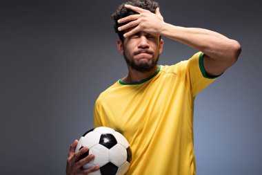 Endişeli bir Afrikalı Amerikan futbol fanatiği sarı tişörtlü ve gri topunu tutuyor.
