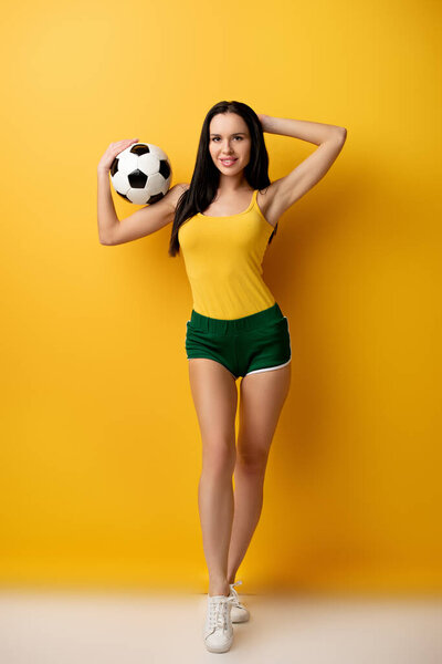 Улыбающаяся футбольная фанатка в шортах держит мяч на желтом
