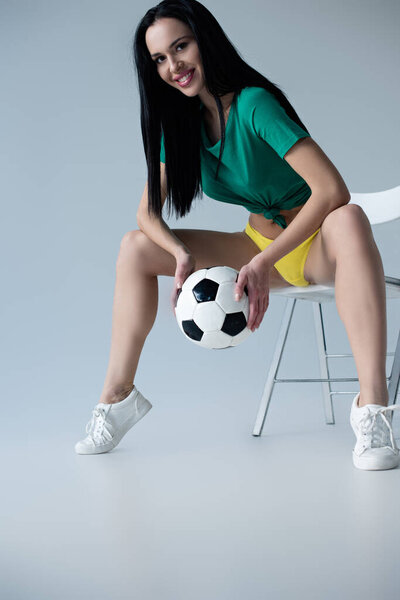 сексуальная улыбающаяся женщина, держащая футбольный мяч, сидя на стуле на сером
