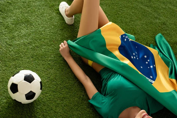 cropped view of sexy girl lying with brazilian flag and football ball on green grass