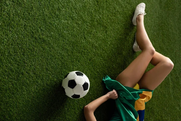 cropped view of sexy girl lying with brazilian flag and football ball on green grass