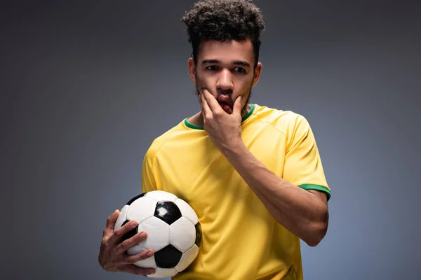 frustrated african american football fan in yellow t-shirt holding ball on grey