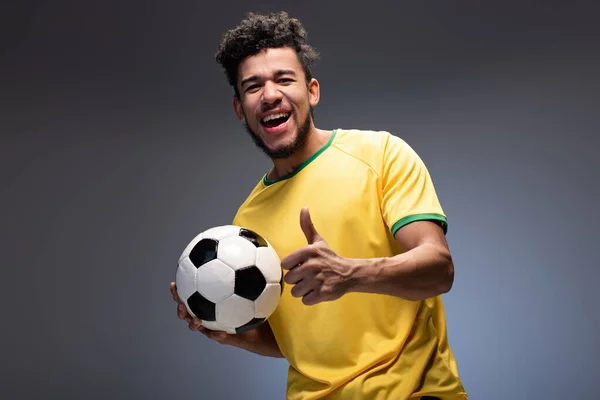 幸せアフリカ系アメリカ人サッカーファン黄色のTシャツを保持ボールとグレーに親指を表示 — ストック写真