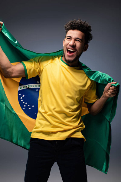 возбужденный африканский американский футбольный болельщик с бразильским флагом на сером
