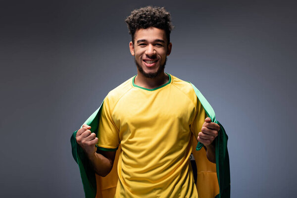 Веселый африканский футбольный болельщик с бразильским флагом на сером
