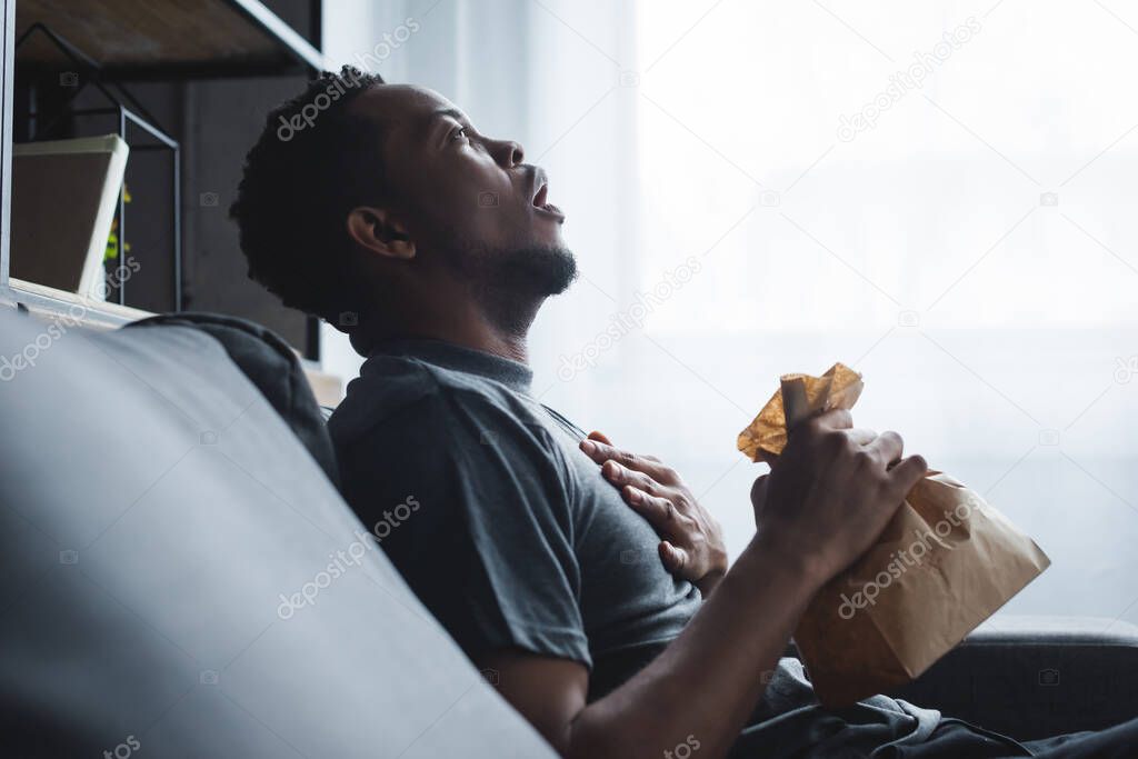shocked african american man holding paper bag while having panic attack at home