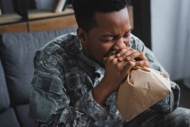 Stresli Afro-Amerikan askeri üniformalı. Evde panik atak ve travma sonrası stres bozukluğundan muzdaripken elinde kese kağıdıyla nefes alıyor.