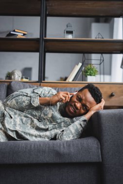 Stresli Afro-Amerikan askeri üniformalı asker akıllı telefonla konuşurken ağlıyor ve evde travma sonrası stres bozukluğu yaşıyor.