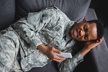 Evde travma sonrası stres bozukluğundan muzdarip, elinde ordu rozeti ve akıllı telefonuyla ağlayan yalnız Afrikalı Amerikan askeri.