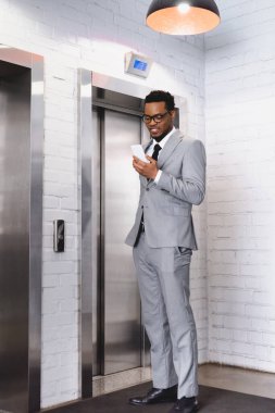 Mutlu Afro-Amerikan iş adamı asansörün yanında dururken akıllı telefon tutuyor.